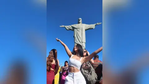 Alison smiles with her arms outstretched in front of the Christ the Redeemer statue wearing a white crop top and white skirt.