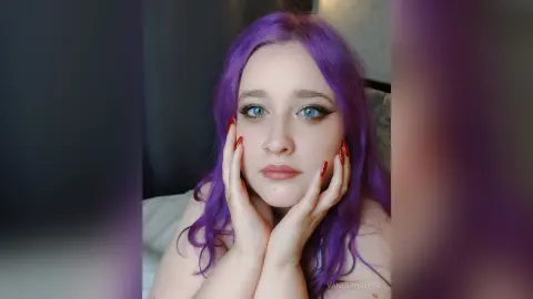 Young woman with purple hair and winged eyeliner posing with hands on face in a soft indoor setting.