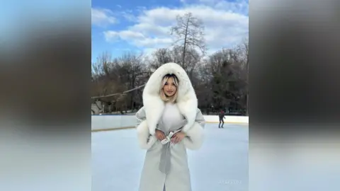 Blonde woman in a white faux fur hooded coat standing on an outdoor ice rink on a sunny winter day.