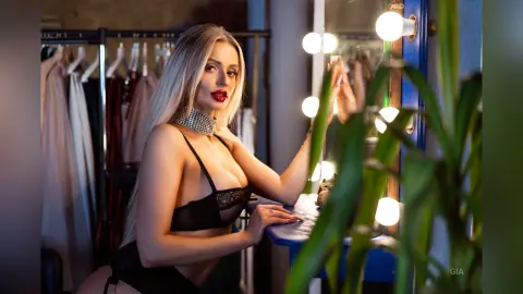 Gia poses in a black lace bra and choker, leaning toward a lit vanity mirror in a dressing room.