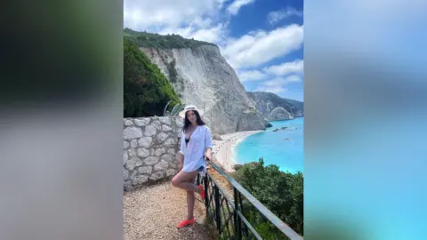 Young woman in a white summer outfit and sun hat posing by a scenic coastal cliff overlooking turquoise ocean water.