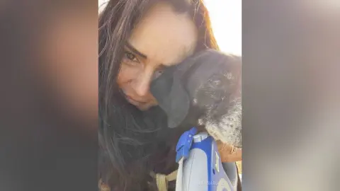 Smiling woman with long dark hair cuddling a spotted grey dog in warm, natural sunlight.