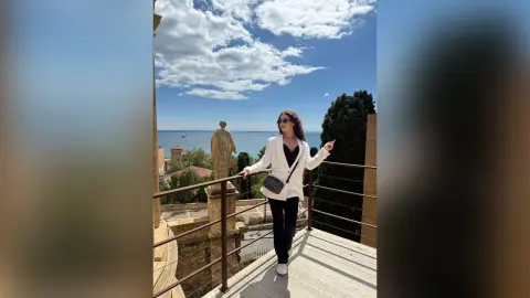 Stylish woman in a white blazer and black trousers posing on a sunny balcony overlooking a coastal town.