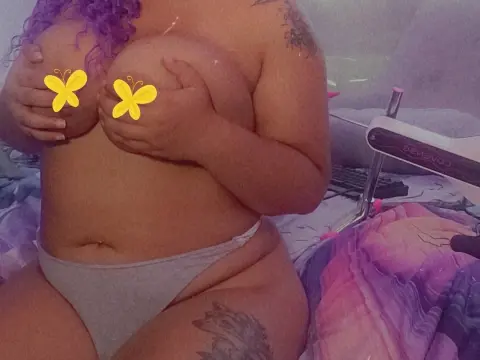 Curvy woman with purple hair in grey bikini bottoms relaxing on a patterned bed in a soft, dreamy mood.