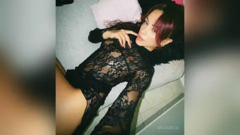 MilaGreco lies on a bed wearing a black lace long-sleeve bodysuit that shows her skin through the floral patterns.
