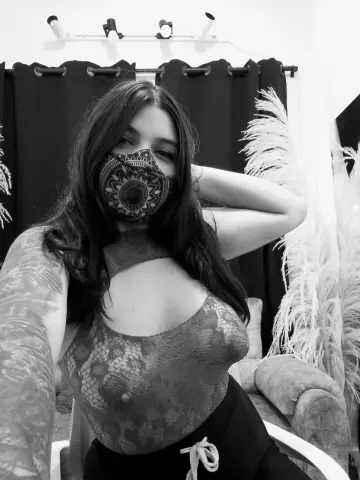 AniFoxx wears a patterned face mask and a sheer lace bodysuit while posing in her studio.