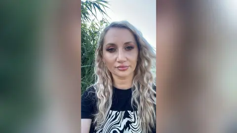 Blonde woman in a black graphic t-shirt smiling outdoors against a lush green nature background.
