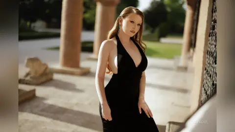 RubySoul poses outdoors in a low-cut black dress, showing off her long red hair and cleavage against a backdrop of stone pillars.