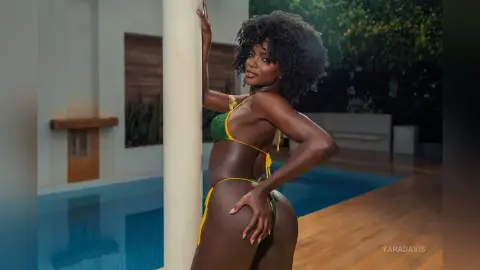 Smiling Black woman in a green bikini posing by a luxury swimming pool at night.