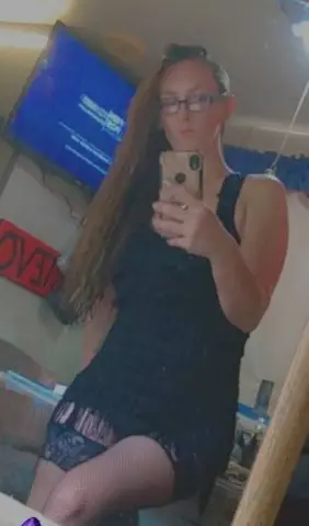 Young woman with long hair in a black sleeveless dress taking a mirror selfie in a cozy room.