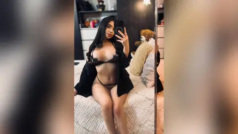 Young woman in black lingerie and an open shirt taking a mirror selfie in a cozy bedroom setting.