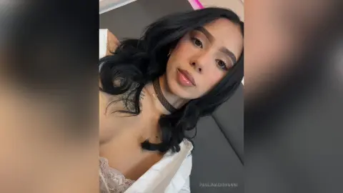 Young woman with long black hair wearing a white lace top and black choker, posing with a soft, moody expression.