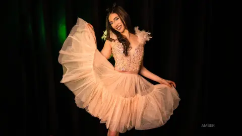Smiling woman in a sparkling blush pink tulle gown posing against a dark background.