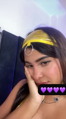 Young woman with long dark hair wearing a yellow headband and gold necklace in a relaxed indoor setting.