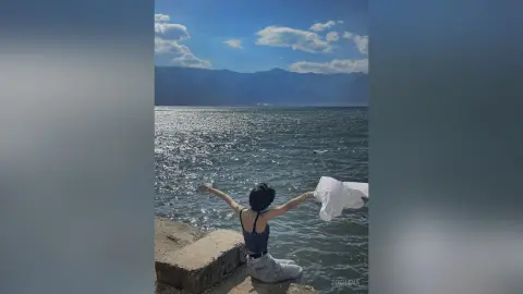 Young woman in a grey tank top sitting by a sparkling blue lake under a sunny sky, enjoying a peaceful moment.