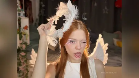 Redheaded woman in white angel costume and feathered headpiece posing playfully in a cozy bedroom setting.