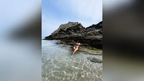 Young woman in a black bikini relaxing in clear ocean water by a rocky coastal cliff under a bright sky.