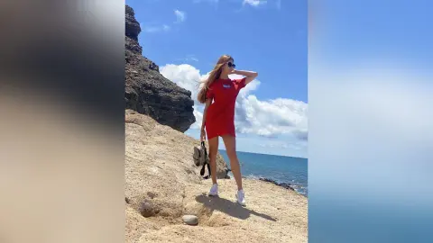 Stylish woman in a red t-shirt dress standing on a rocky cliff overlooking the blue ocean under a sunny sky.