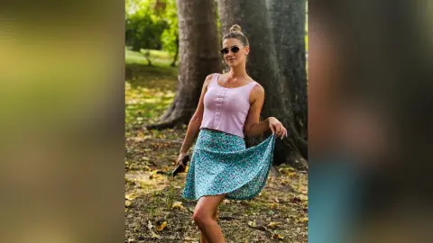 Stylish woman in a pink tank top and floral teal skirt posing in a sunny park setting.