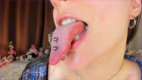 Close-up of a woman with a tongue piercing and silver hoop earring smiling in a soft, natural setting.