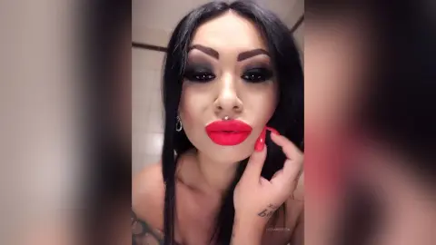 DeeaAnderson poses with long black hair and bright red lipstick, touching her face with a red-nailed hand.