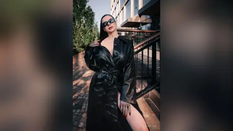 Stylish woman in a black leather trench coat and sunglasses posing confidently on a city street.