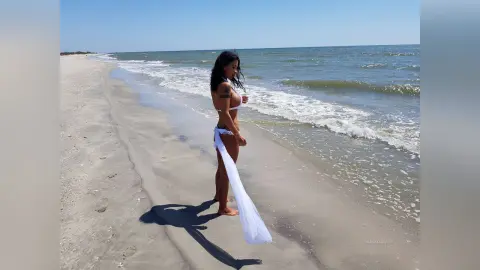 Woman in a white bikini and flowing sarong walking along a sunny beach with ocean waves.