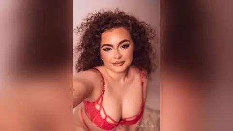 Young woman with curly hair wearing a red lace bra in a warm, intimate indoor setting.