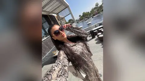 Stylish woman in snakeskin print blouse wearing sunglasses at a sunny outdoor waterfront cafe.