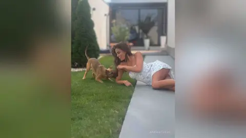 Smiling woman in a white floral dress playing with a brown dog on a sunny patio garden.
