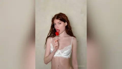 Redheaded woman in white lace lingerie eating a red lollipop against a neutral textured wall.