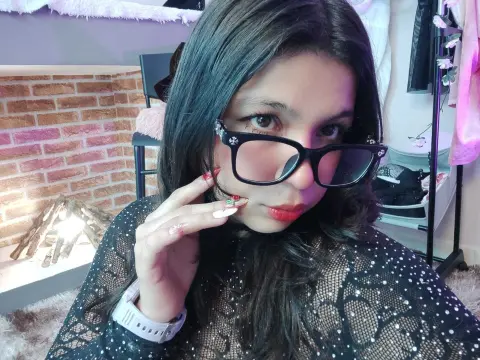 Young woman with long dark hair wearing black glasses and a black sequin lace top in a cozy room.