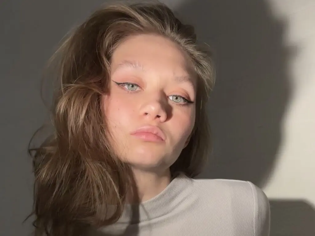 Young woman with winged eyeliner and wavy hair wearing a white ribbed turtleneck in soft studio lighting.