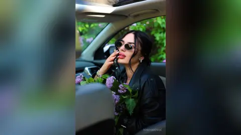 Stylish woman in a black leather jacket and sunglasses holding purple flowers inside a car.