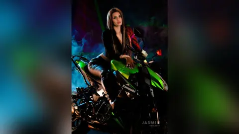 AndreaKalo poses on a green Kawasaki motorcycle, wearing a black long-sleeved top and black leggings or boots, surrounded by smoke.