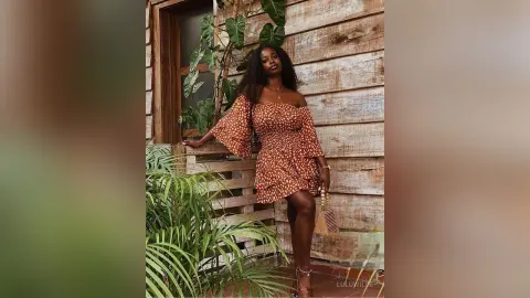 Stylish Black woman in a brown floral off-the-shoulder mini dress posing on a rustic wooden balcony.