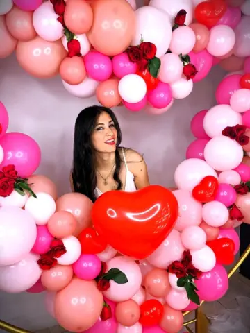 Smiling woman in a white top posing within a festive pink and red balloon arch for a romantic celebration.