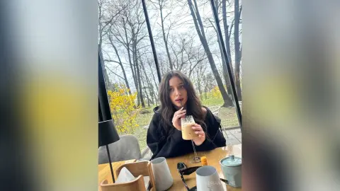 Young woman in a black sweater sipping iced coffee at an outdoor cafe on a cloudy day.