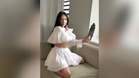 TessaWinsent — TessaWinsent sits on a beige sofa wearing a white ruffled crop top and matching mini skirt while holding a mirror.