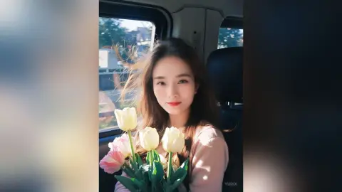 Smiling young woman in a pink sweater holding a bouquet of white tulips inside a car on a sunny day.