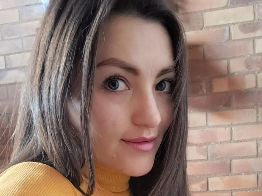 Smiling young woman with long brown hair wearing a yellow ribbed turtleneck against a brick wall background.