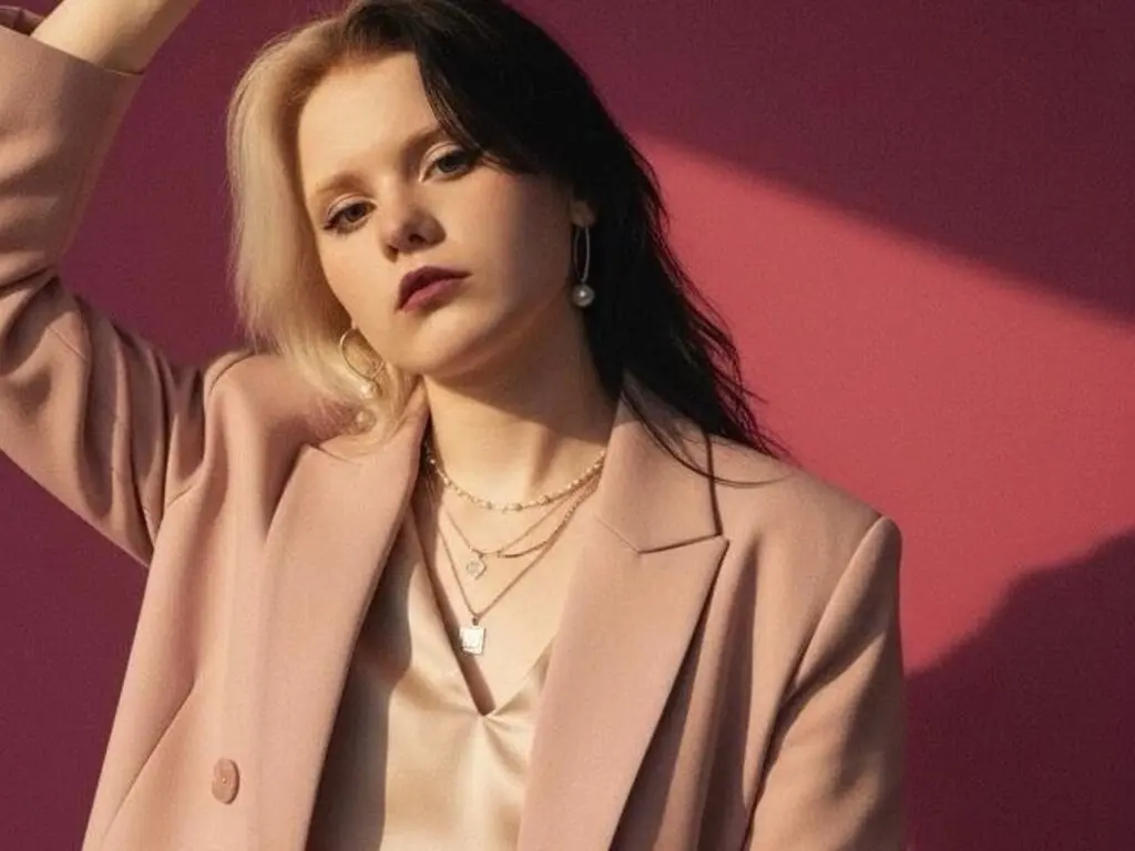 Stylish young woman in a beige blazer and gold jewelry posing against a pink background in warm sunlight.