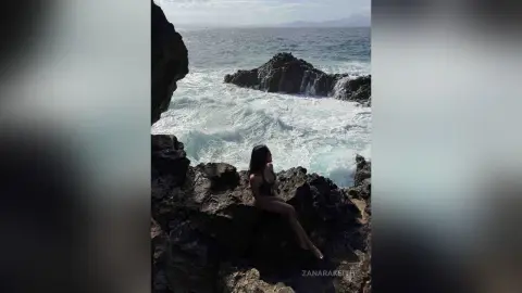 Woman in a black swimsuit sitting on dark coastal rocks overlooking crashing ocean waves under a bright sky.