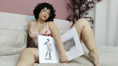 Young woman with curly hair in a floral camisole relaxing on a bed while reading an art book.