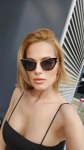 Blonde woman in a black camisole and dark sunglasses posing indoors with a modern aesthetic.