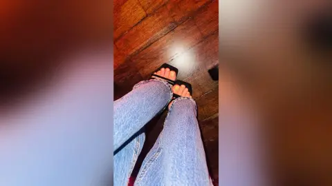 Close-up of a person wearing light wash denim jeans and black platform slide sandals on a dark wood floor.