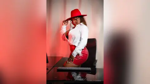 Stylish woman in a red hat and white blouse sitting at a desk in a moody, red-lit studio setting.