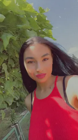 Young woman in a red ribbed tank top smiling outdoors against a lush green leafy background.