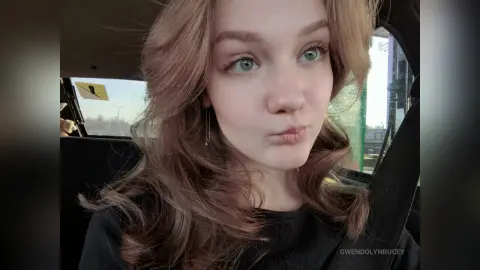 GwendolynBucey pouts her lips while looking off-camera, showing her long wavy blonde hair and wearing a black top while sitting in a car.