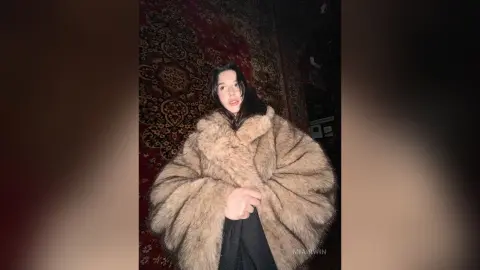 Young woman in a cozy oversized faux fur coat posing against a vintage patterned rug in moody lighting.